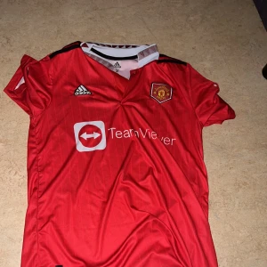 Manchester United Ronaldo 7 tröja - Säljer en officiell Manchester United fotbollströja från Adidas med Ronaldo och nummer 7 på ryggen. Tröjan är röd med tunna vertikala ränder, korta ärmar och klubbmärke på bröstet. Materialet är lätt och andas, perfekt för match eller träning.