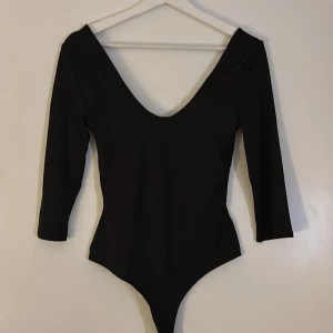 Svart långärmad bodysuit från H&M - Svart bodysuit från H&M med djup v-ringning och lång ärm. Snygg, tight passform som framhäver siluetten. Tillverkad i mjuk och stretchig polyester som sitter bekvämt mot kroppen. Perfekt för att styla med jeans eller kjol.