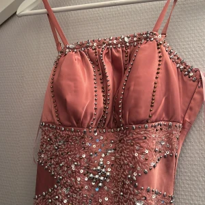 Rosa långklänning med paljetter och pärlor - Säljer en glittrig rosa långklänning med smala axelband och vackra paljett- och pärldetaljer upptill. Klänningen har omlott-snörning i ryggen och är tillverkad i ett glansigt material som ger en lyxig känsla. Perfekt för speciella tillfällen!