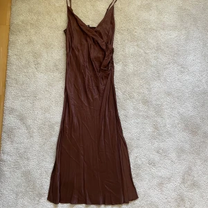 Brun omlottklänning med smala axelband L oanvänd  - Säljer en lång brun klänning med smala axelband och omlottdetalj framtill. Klänningen har en elegant v-ringning och är tillverkad i ett mjukt, glansigt material som ger en lyxig känsla. Perfekt för dig som gillar stilrena och enkla plagg med en twist.