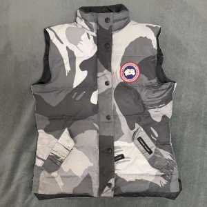 Canada Goose väst  - Säljer en snygg dunväst från Canada Goose i grått och vitt camouflage-mönster. Västen har hög krage, tryckknappar och dragkedja framtill samt två fickor med lock. Perfekt för lager-på-lager-stil.