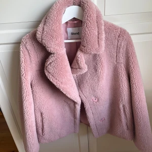 Rosa teddyjacka från Stand - Supermjuk och fluffig rosa teddyjacka från Stand med bred krage och knappar framtill. Jackan har en klassisk, lite boxig passform och är perfekt för kyligare dagar. Fodrad insida för extra komfort.