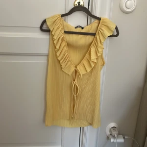 Gul volangprydd ärmlös topp från Zara - Säljer en ljusgul ärmlös topp från Zara med djup v-ringning och volangkant längs ringningen. Toppen har knytband framtill och är tillverkad i ett lätt, plisserat material. Perfekt för vår och sommar!