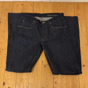 Mörkblå jeans från Dunderdon - Säljer ett par klassiska mörkblå jeans från Dunderdon med raka ben och orange kontrastsömmar. Jeansen har fem fickor, knappgylf och är tillverkade i kraftigt denimtyg. Perfekta för dig som gillar stilrena och tidlösa jeans.