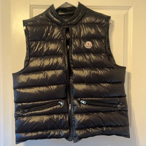 Svart dunväst från Moncler - Snygg svart dunväst från Moncler med hög krage och två dragkedjefickor framtill. Västen har ett klassiskt Moncler-märke på bröstet och stängs med dragkedja. Äkta.