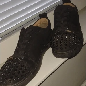 Svarta sneakers med nitar och röd sula från Christian Louboutin - Säljer ett par svarta sneakers från Christian Louboutin med ikonisk röd sula och nitar framtill. Skorna har snörning och är tillverkade i mocka. Perfekta för dig som vill sticka ut med en edgy stil.