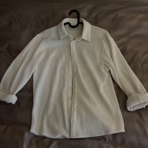 Vit ribbad skjorta från Grunt - Säljer en vit skjorta från Grunt i storlek L i barn storlek(ungefär xs i herr storlek). Skjortan har ribbad struktur, klassisk krage och knäppning framtill. Perfekt för dig som gillar en stilren och enkel look i sommar. Långa ärmar och mjukt material.
