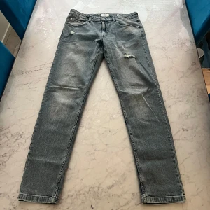Grå slitna jeans från Rebel - Säljer ett par grå jeans från Rebel med slitningar och hål på ena benet. Klassisk femficksmodell med dragkedja och knapp. Raka ben och normal passform. Storlek W34 L34.