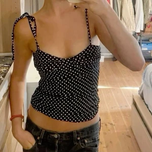 Svart prickig linnetopp från Zara - Supersöt svart body från Zara med vita prickar och smala axelband som knyts på axlarna. Toppen har en figurnära passform och lätt rynkade detaljer framtill. Perfekt för varma dagar och enkel att styla med jeans eller kjol.