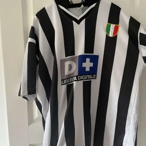 Juventus fotbollströja Zidane 21 - Klassisk Juventus fotbollströja med svarta och vita ränder, korta ärmar och V-ringning. Tryck med 'Zidane' och nummer 21 på ryggen, klubbmärke på ärmen och italienskt mästerskapsmärke på bröstet. Sponsorlogga 'Liberta Digitale' på framsidan.