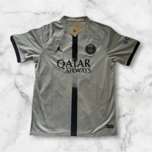 PSG fotbollströja Jordan x Qatar Airways - Snygg PSG fotbollströja i silvergrått med svart vertikal rand och svarta detaljer vid ärmslut. Jordan-logga och PSG-emblem på bröstet samt Qatar Airways tryck på framsidan. Tillverkad i lätt och ventilerande material.