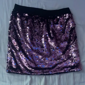 Rosa paljettkjol - Glittrig rosa paljettkjol från Lindex med svart midjeresår. Kjolen är medellång och täckt av skimrande paljetter som reflekterar ljuset i lila och rosa toner. Perfekt för dig som vill sticka ut och glänsa eller till en skolbal.