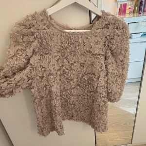 Beige spetsblus med 3D-blommor från Holly & Whyte - Supersöt beige blus från Holly & Whyte med 3D-blommor och spetsdetaljer. Blusen har rund halsringning och långa ärmar. Perfekt för dig som vill sticka ut med en romantisk look.