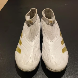 Adidas Predator fotbollsskor i vitt och guld - Säljer ett par Adidas Predator fotbollsskor i vitt med guldiga detaljer. Skorna har hög strumpliknande krage och är utan snörning för en tight passform. Perfekta för spel på gräsplaner med fasta dobbar och snygg design.