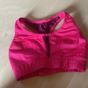 Rosa sport-bh från Gina Tricot - Säljer en starkt rosa sport-bh från Gina Tricot med brottarrygg och justerbar knäppning bak. Bh:n har bred resår nedtill och är tillverkad i ett stretchigt syntetmaterial. Perfekt för träning och ger bra stöd.