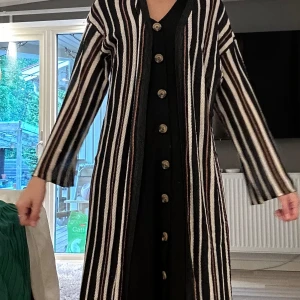 Svart och vit kofta  - Säljer en lång kofta med svarta, vita och beige vertikala ränder. Koftan har långa ärmar och stora knappar framtill. 