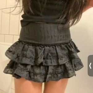 Svart volangkjol med broderi 38 - Supersöt svart kortkjol med flera volanger och broderade detaljer. Hög midja och figurnära passform upptill, med broderat mönster och lätt rynkade volanger som ger kjolen extra volym och en trendig look.