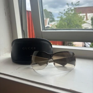 Solglasögon från Gucci - Snygga solglasögon från Gucci med gråtonade linser och beige skalmar dekorerade med Guccis logomönster. Kommer med originalfodral. Perfekta för att ge en lyxig touch till din stil.