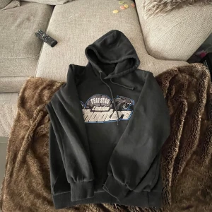 Svart hoodie från Trapstar - Säljer en svart hoodie från Trapstar med tryck på bröstet i vitt och blått. Tröjan har huva med dragsko och ribbade muddar vid ärmslut och nederkant. Perfekt för en avslappnad och cool stil.