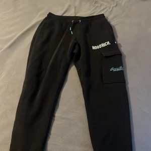 Svarta mjukisbyxor från Hoodrich - Svarta mjukisbyxor från Hoodrich med tryckt logga på benet och textdetaljer. Byxorna har dragsko i midjan och en ficka med lock på sidan. Perfekta för en avslappnad stil och streetwear-vibe. Använder dom inte då dom e för små för mig.