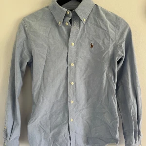 Ralph Lauren skjorta  - Klassisk ljusblå skjorta från Polo Ralph Lauren med broderad logga på bröstet. Skjortan har knappar hela vägen, button-down krage och långa ärmar. Perfekt för alla situationer. Skjortan har extremt bra skick utan några skador. Hör av dig för mer frågor och funderingar!