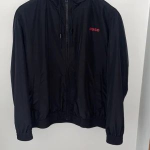 Svart vindjacka från Hugo - Svart vindjacka från Hugo med röd logga på bröstet och röd meshfoder på insidan av huvan. Jackan har dragkedja framtill, huva med dragsko och elastiska muddar vid ärmslut och midja. Perfekt för blåsiga dagar.