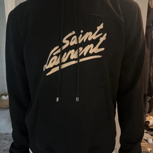 Svart hoodie från Saint Laurent - Säljer en svart hoodie från Saint Laurent med stor vit logga framtill tröjan är M men passar S. Tröjan har huva med dragsko och en stor magficka. Perfekt för dig som gillar stilrena och exklusiva plagg. Skriv om frågor , kan bytas 
