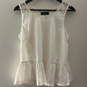 Vit ärmlös blus med spetsdetaljer - Säljer en vit ärmlös blus från Sister S point med spetsdetaljer på ryggen. Blusen har en volang nertill och är tillverkad i ett lätt, glansigt material. Sitter som s/m. Skriv för fler bilder!