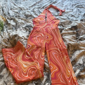 Färgglad byxdress med virvlande mönster - Säljer en snygg byxdress med virvlande mönster i rosa, orange och vitt. Byxdressen har halterneck med knyt i nacken och vida ben. Perfekt för dig som vill sticka ut med färg och form!