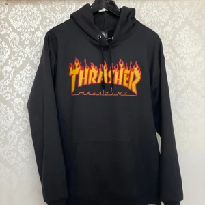 Svart Thrasher hoodie med flames - Säljer en svart hoodie från Thrasher med den klassiska loggan i gult och rött med flames på bröstet. Tröjan har huva med dragsko och en stor magficka. Perfekt för dig som gillar streetwear och skate.