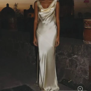 Champagnefärgad långklänning i satin - Elegant långklänning i skimrande champagnefärgad satin med draperad urringning och öppen rygg. Klänningen har smala axelband och en figurnära passform som ger en lyxig känsla. Perfekt för speciella tillfällen. Säljer då den sitter för tajt runt vissa ställen.. De ska va S men skulle säga att de mer är XS för dem med större byst