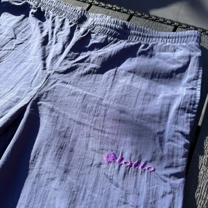 Ljuslila träningsbyxor från Lotto - Lätta och luftiga ljuslila byxor från Lotto med resår i midjan och raka ben. Byxorna har broderad logga i lila och rosa på ena benet. Perfekta för varma dagar och avslappnad stil.