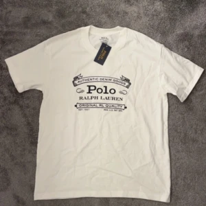 Vit t-shirt från Polo Ralph Lauren - Vit t-shirt från Polo Ralph Lauren med svart tryck på bröstet. Modellen är custom slim fit och har rund halsringning. Perfekt för en stilren och klassisk look. Tillverkad i mjuk bomull.