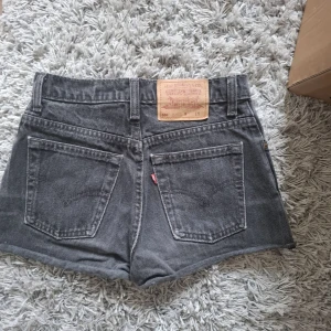 Grå jeansshorts från Levi's - Säljer ett par grå jeansshorts från Levi's med klassisk femficksdesign och råa kanter nertill. Shortsen har hög midja och synliga sömmar, perfekt för en avslappnad stil.