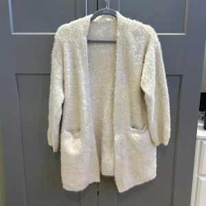 Beige fluffig kofta från Women'secret - Mysig och fluffig beige lång kofta från Women'secret med långa ärmar och två stora fickor framtill i fluffigt material. I strl M