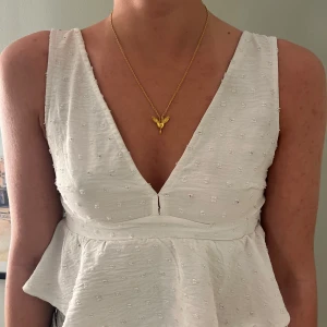 Vit ärmlös blus med volang - Säljer en söt vit blus med breda axelband och volang nertill. Blusen har diskret strukturmönster och djup v-ringning framtill. Dragkedja i ryggen för enkel påklädning. Perfekt för varma dagar!