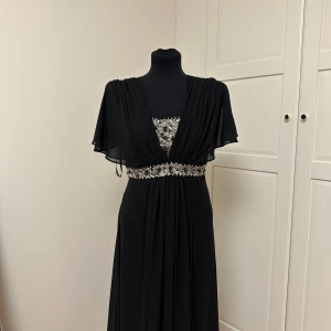 Svart långklänning med glittriga detaljer - Elegant svart långklänning med v-ringning och glittriga paljettdetaljer vid midjan och bysten. Klänningen har korta, fladdriga ärmar och ett mjukt, fallande tyg som ger en lyxig känsla! Storleken är L och klänningen är endast använt 1 gång så den är i mycket god skick! 