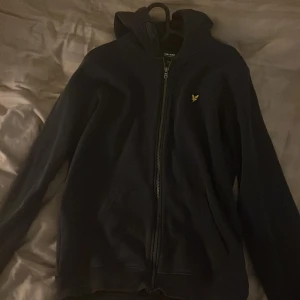 mörkblå  hoodie från Lyle & Scott Junior - Mörkblå hoodie från Lyle & Scott Junior med dragkedja och huva. Jackan har det klassiska gula logobroderiet på bröstet och två fickor framtill. Perfekt för en avslappnad stil. står 15/16 men tycker själv den passar som xs