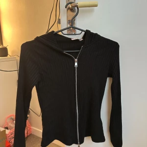 Svart ribbad hoodie med dragkedja - Svart ribbad hoodie med dragkedja framtill och huva. Jackan har lång ärm och är i ett stretchigt material som sitter tajt mot kroppen. Perfekt för lager-på-lager och enkel att matcha med olika outfits. Helt ny 