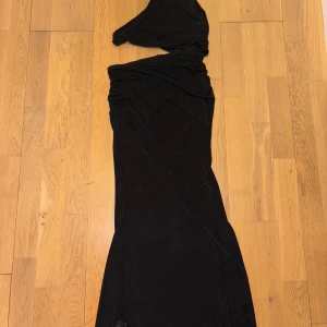 Svart långklänning med oneshoulder - Snygg svart långklänning med oneshoulder-design och draperade detaljer upptill. Klänningen har en elegant och smickrande passform som ger en cool och sofistikerad vibe. Perfekt för dig som vill sticka ut med en stilren look.