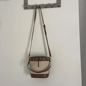 Beige och brun bucket bag med snörning från TEMU - Snygg bucket bag i beige canvas med bruna skinn-detaljer och guldfärgade metalldelar. Väskan har både axelrem och handtag, samt snörning upptill. Insidan är fodrad med randigt tyg och har en innerficka med dragkedja. Perfekt för dig som vill ha en stilren och trendig väska. (Aldrig använd)