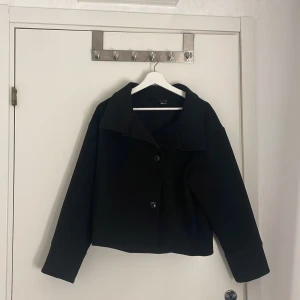 Svart kappa från Gina Tricot XS - Snygg svart kappa från Gina Tricot i kortare modell med bred krage och knäppning framtill samt två stora fickor. Jackan har raka, långa ärmar och en stilren look som passar perfekt till höst och vinter. 
