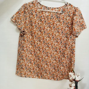Blommig kortärmad blus i orange och beige - Söt blus med småblommigt mönster i orange, beige och brunt. Modellen är rak med rund halsringning och korta ärmar. Perfekt för dig som gillar en romantisk och avslappnad stil.