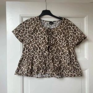 Leopardmönstrad blus från Gina Tricot - Superfin blus från Gina Tricot i leopardmönster med bruna, svarta och beige toner. Modellen är luftig med korta ärmar, volang nertill och snörning framtill. Perfekt för dig som vill sticka ut med ett djurmönstrat plagg.