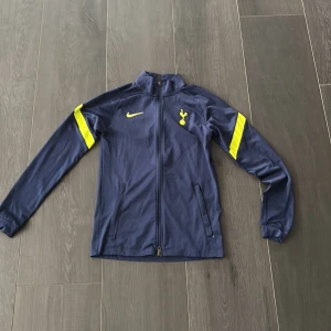 Tottenham Nike blå vindjacka - Snygg marinblå vindjacka från Nike med Tottenham Hotspur-logga och gula detaljer på bröstet och ärmarna. Jackan har hel dragkedja framtill, ståkrage och två sidofickor. Perfekt för dig som gillar fotboll och vill ha en sportig stil.