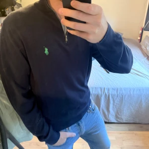 Marinblå half zip tröja Polo Golf - Säljer en marinblå långärmad tröja från Polo Golf Ralph Lauren i storlek XL. Tröjan har en klassisk half zip-dragkedja och grönt broderat logotyp på bröstet. Ribbstickade muddar och krage med grön rand på insidan. Perfekt för dig som gillar stilren och sportig look.passar liten, liknar storlek L
