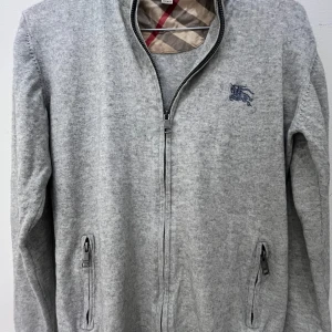 Grå kofta från Burberry med dragkedja - KAN GÅ NED I PRIS VID SNABB AFFÄR! Stilren grå kofta från Burberry med klassiskt rutigt foder i kragen och broderad logga på bröstet. Långärmad modell med dragkedja framtill och fickor med dragkedja. Perfekt för dig som gillar stilrena och exklusiva plagg. Ny skick! Skulle säga att den passar allt mellan 160-167 perfekt!