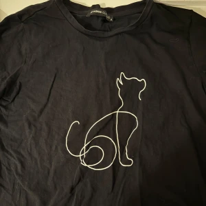 Svart t-shirt med kattmotiv  - Svart t-shirt med ett stilrent vitt kattmotiv på framsidan. Klassisk rund hals och korta ärmar. Materialet är mjuk bomull som känns skönt mot huden. Perfekt för dig som gillar minimalistiska prints och djurmotiv.