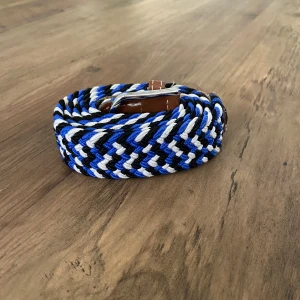 RYVLS flätat bälte med läderdetaljer - Snyggt flätat bälte i blått, svart och vitt med bruna läderdetaljer och silverspänne. Bältet har ett randigt mönster och passar perfekt till jeans eller chinos för en avslappnad stil. Justerbar längd tack vare det flexibla materialet.