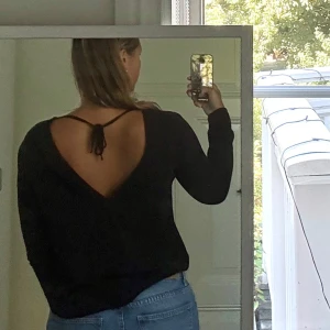 Svart blus med öppen rygg  - Svart blus från NLY Trend i storlek 38. Blusen har lång ärm med knapp vid ärmslut, djup v-ringad öppen rygg med knytband upptill. Tunt, lätt strukturerat material som känns luftigt.